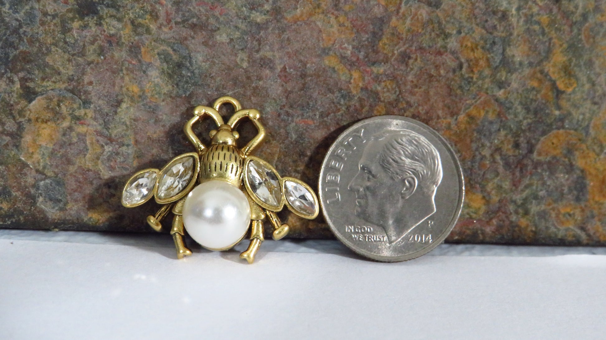 Gold bee-shaped brooch with pearls next to a quarter on a textured surface