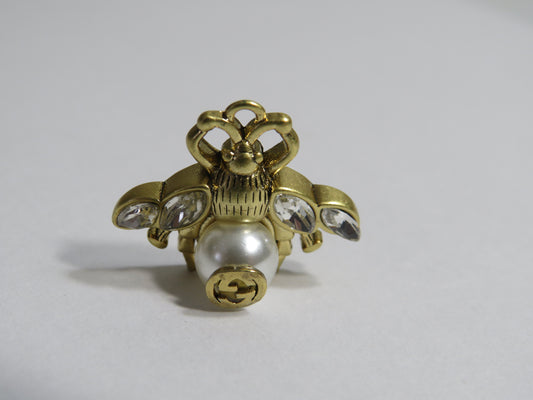 A vintage-style gold-tone ring shaped like a bee, featuring a white faux-pearl bead on the abdomen, clear rhinestones on the wings, and a Gucci logo on the belly, set against a plain light gray background.